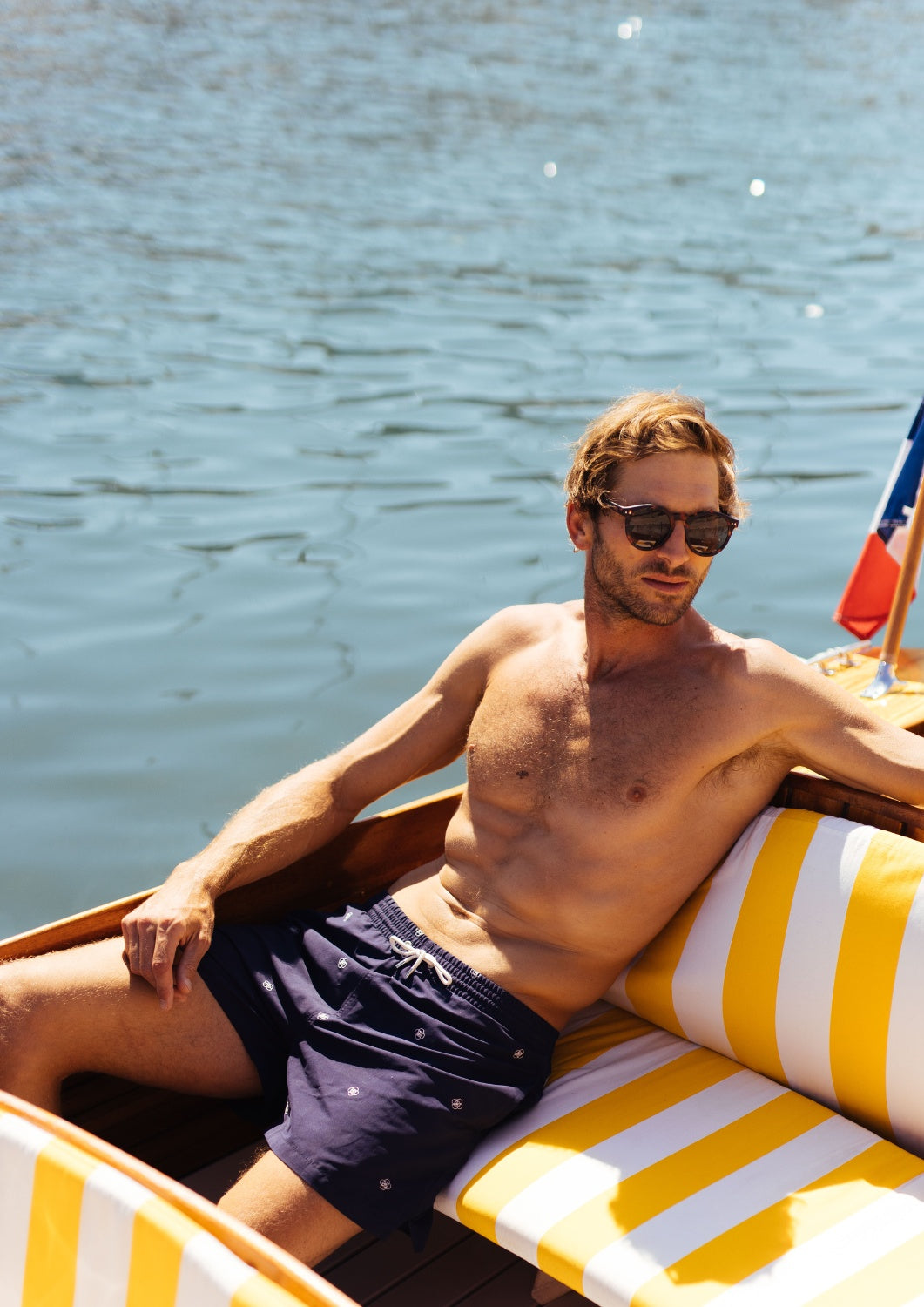short maillot de bain homme porté par un bel homme blond au soleil dans un bateau avec des tissus rayés jaunes