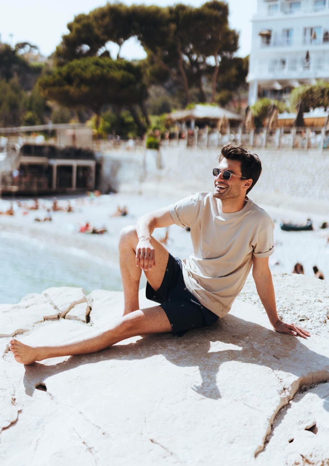 short maillot de bain homme porté par un homme avec un haut beige et des lunettes de soleil proche de la plage